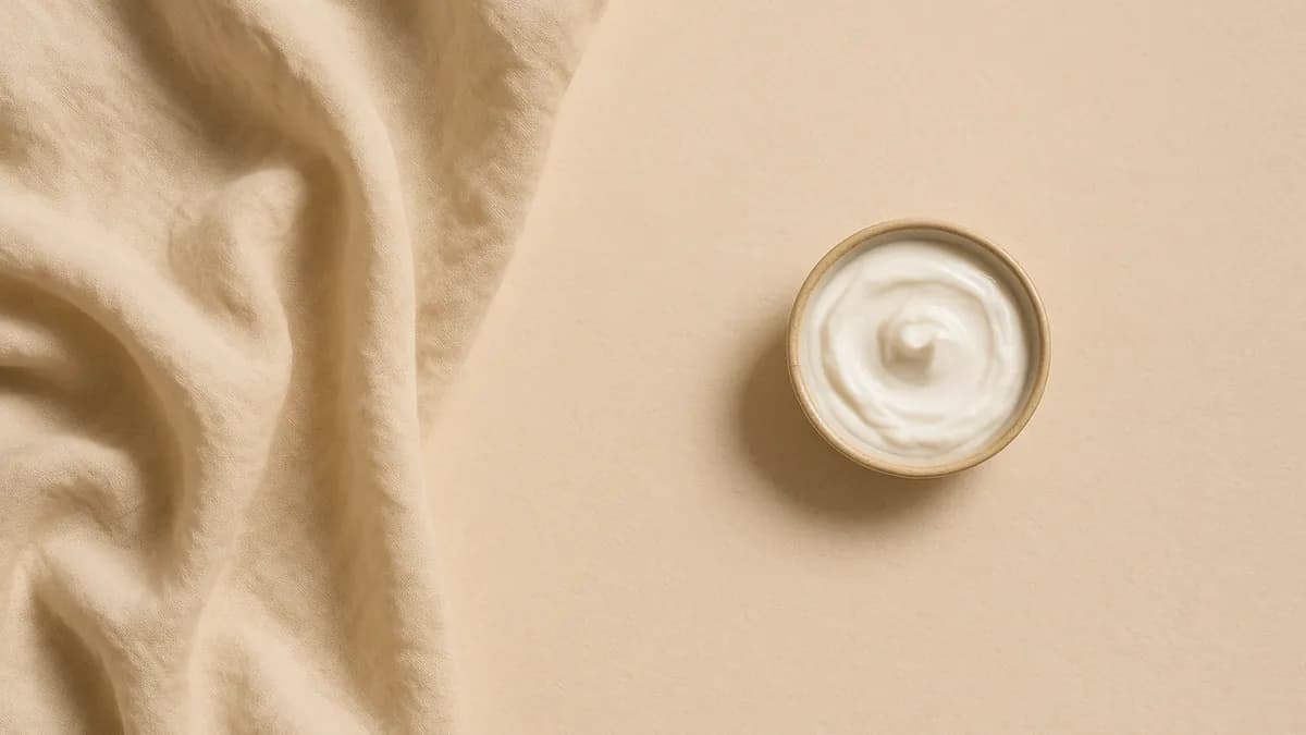 Soft cream linen in loose folds beside a small dish of white body cream